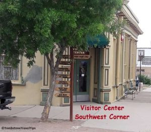 Tombstone Visitor Center Entry, tree on corner & directional arrow signs for town attractions. Bench around the corner by the Tombstone Art Gallery.